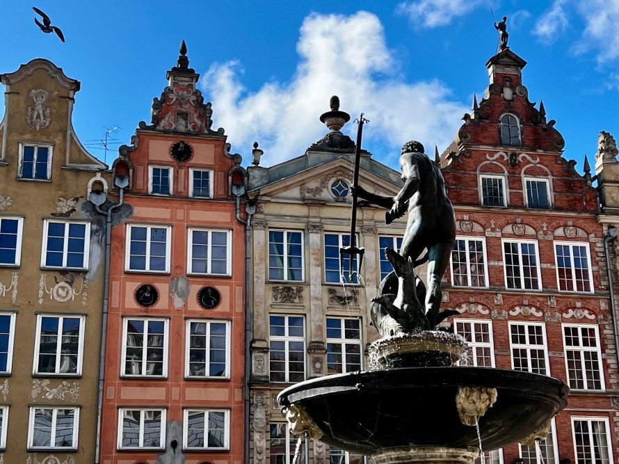 Fuente con una estatua de un hombre sosteniendo un tridente, frente a edificios de estilo arquitectónico clásico en una plaza iluminada por el sol.