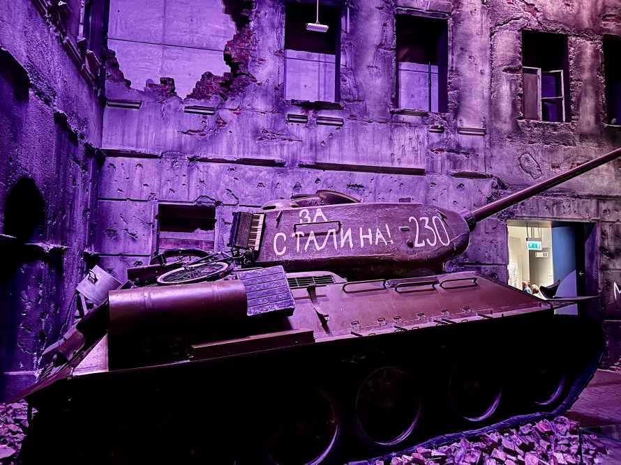 Tanque militar en un edificio en ruinas con grafiti que dice 'ЗА СТАЛИНА! -230'. Iluminación en tonos morados.