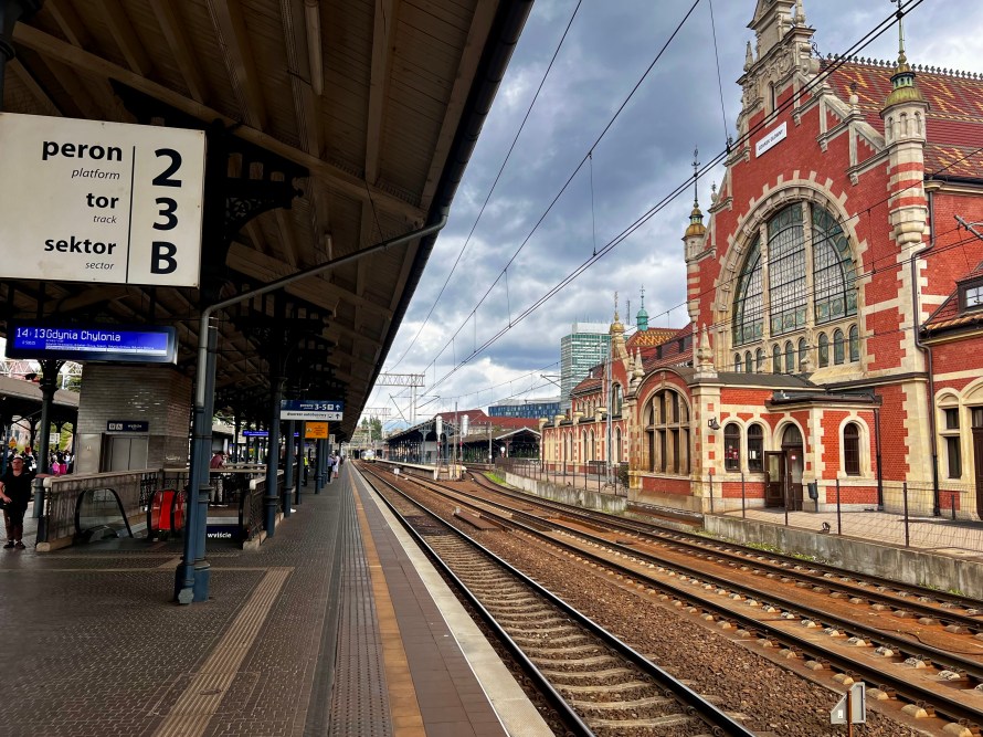 Plataforma 23B de una estación de tren con un edificio histórico al fondo y vías de tren visibles.