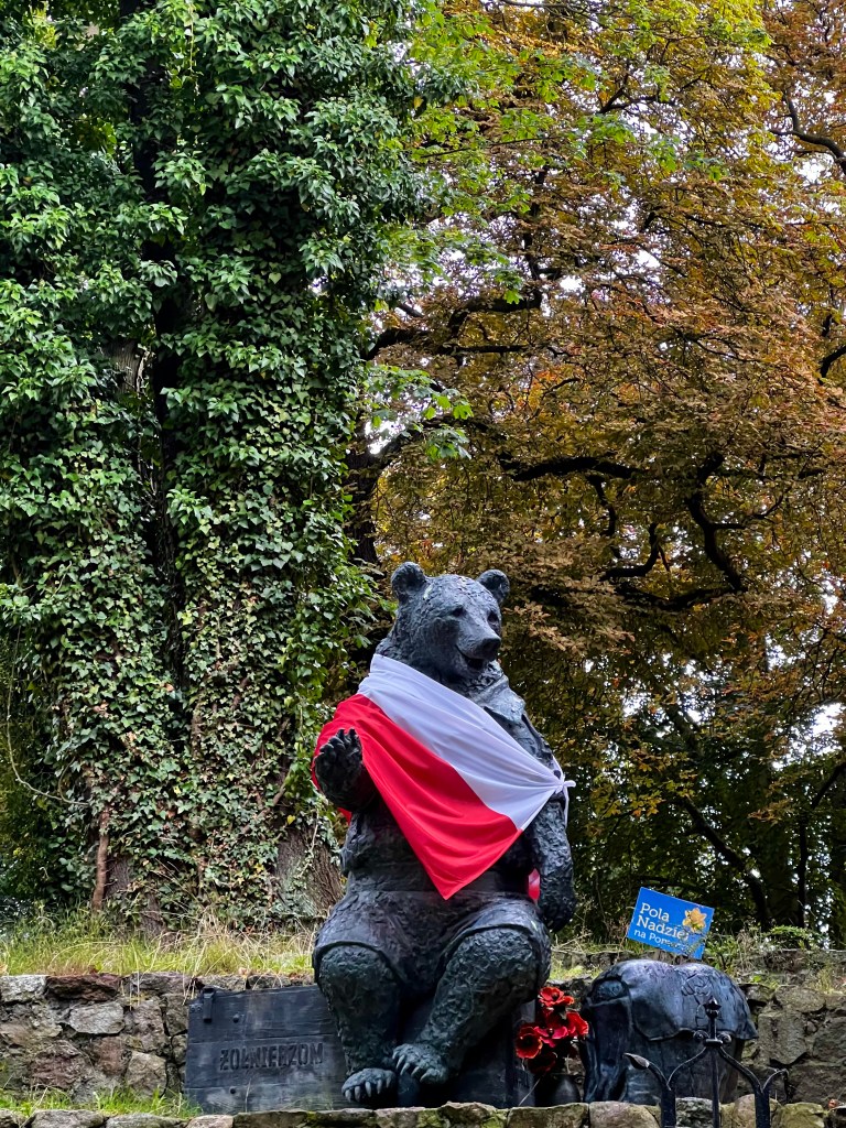 Estatua de un oso con una bandera polaca y flores en un entorno natural con árboles.