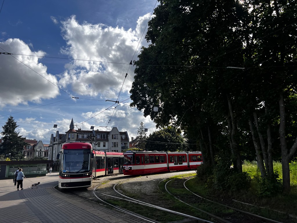 Tren ligero en una vía con edificios y árboles al fondo bajo un cielo nublado.