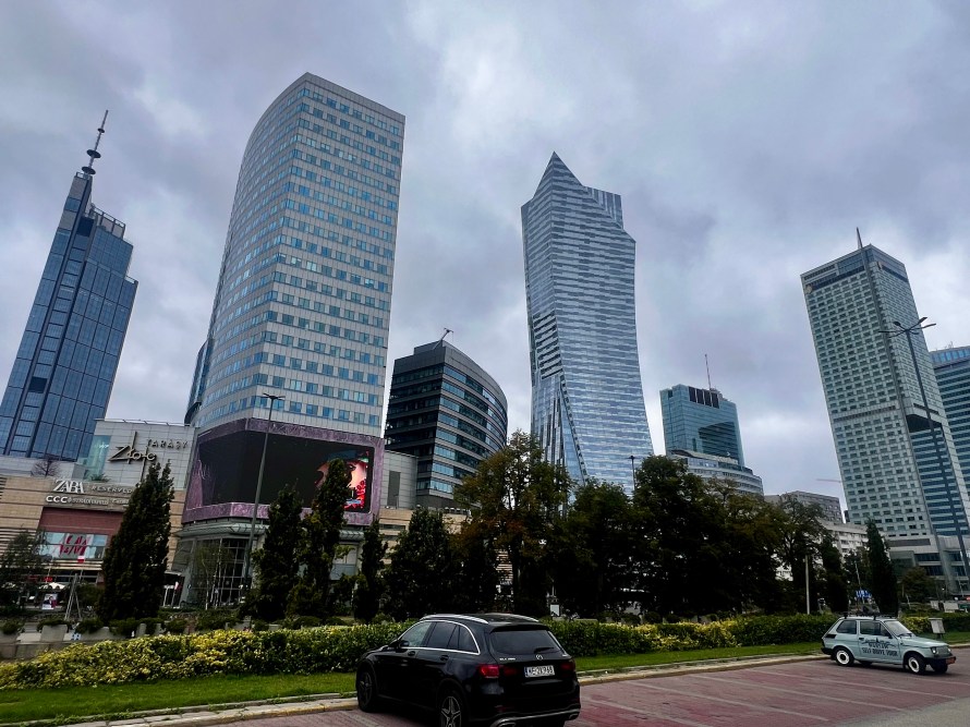 Vista de un grupo de rascacielos en una ciudad con cielo nublado, incluyendo edificios de vidrio y un coche aparcado en primer plano.